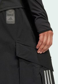 Black athletic jacket featuring a sleek design, textured fabric, adjustable pocket, and three white stripes on the side. Logo patch visible.