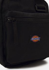 Zwarte stoffen rugzak met een voorvak met rits, stevige schouderbanden en een kleurrijke "Dickies" logo patch aan de zijkant. Duurzaam en functioneel ontwerp.