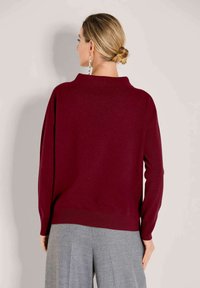 Vrouw met blond haar in een lage knot draagt een ruim vallende bordeauxrode trui en grijze wijde broek, gezien vanuit de rug.