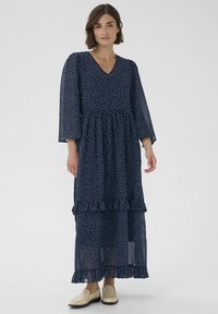 Marineblaues Maxikleid mit weißen Polka-Dots, V-Ausschnitt, ausgestellten Ärmeln, gestuften Rüschen am Saum, aus leichtem, transparentem Stoff gefertigt.