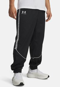 Black track pants made of smooth fabric, featuring white piping and side stripes with a logo. Tapered fit with elastic cuffs at the ankles.