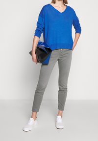 Pull bleu à col en V, coupe décontractée, associé à un jean skinny gris clair et des baskets blanches. Sac à main noir et bleu tenu dans la main droite.