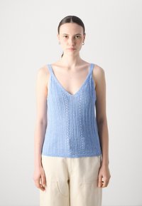 Lyseblå strikket tanktop med V-hals, med et teksturert mønster og tynne skulderstropper. Kombinert med løse, kremfargede bukser.
