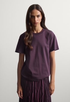 Donna con capelli lunghi e scuri che indossa una maglietta viola semplice a maniche corte e una gonna plissettata viola, in piedi su uno sfondo grigio chiaro.