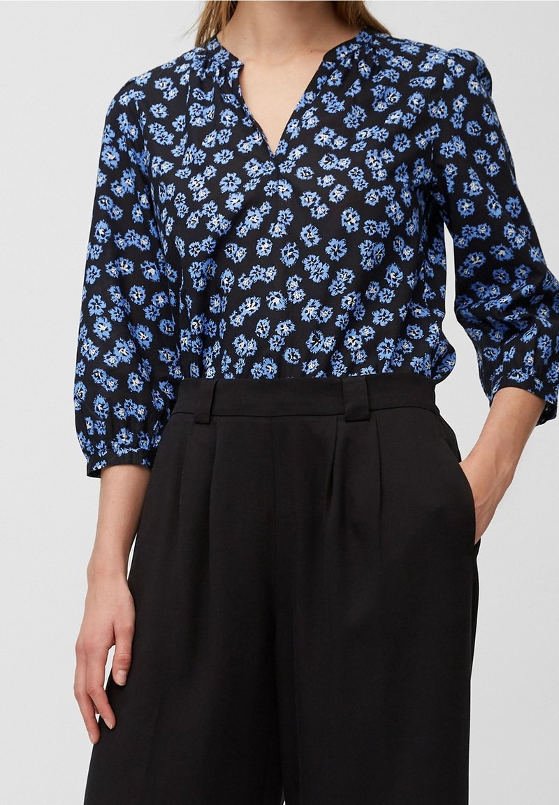 Blusa nera a fiori con fiori blu, scollo a V e maniche a sbuffo a tre quarti, abbinata a pantaloni neri a vita alta.