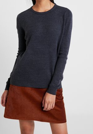 Femme portant un pull à manches longues gris foncé texturé et une jupe courte en velours côtelé couleur rouille, debout devant un fond uni.
