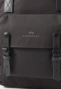 Sac à dos en tissu noir avec des accents en cuir, comportant une poche avant sécurisée par deux sangles. Le nom de la marque "DOUGHNUT" est embossé en doré.