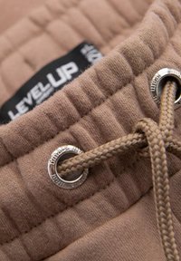 Brauner gesteppter Stoff mit strukturiertem Finish. Verfügt über ein silbernes Metallösen und eine gewebte beige Kordel. Label mit "LEVEL UP" sichtbar.