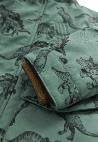 Veste imperméable verte avec un motif de dinosaure noir, dotée d'un poignet texturé souligné de tissu marron et parsemé de gouttes d'eau.