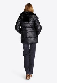 Schwarze Daunenjacke mit einer Kapuze aus Kunstfell, gestepptem Design und einem etwas längeren Schnitt. Kombiniert mit dunklen Jeans und Schuhen mit Tierdruck.