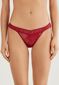 Red lace thong with a sheer mesh body, floral detailing, and a patterned elastic waistband. Smooth texture with delicate trim accents.