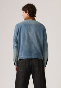 Άνδρας που φαίνεται από πίσω, φορώντας μια μπλε τζιν μπουφάν με καφέ γιακά και μαύρο τζιν παντελόνι, μπροστά σε ένα απλό φωτεινό φόντο.