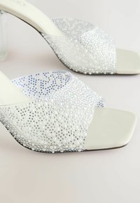 Des sandales à talons hauts blanches avec un dessus en perles iridescentes, agrémenté de petites perles et de strass, ainsi qu'un talon bloc transparent.