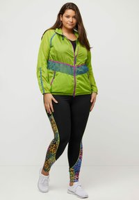 Grön vindjacka med lila detaljer och nätpanel, tillsammans med svarta leggings med flerfärgade blommönster längs sidorna.