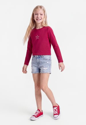 Lachend meisje met lang blond haar draagt een rood shirt met lange mouwen en een ster, lichte gescheurde spijkershorts en rode sneakers, staand op een witte achtergrond.