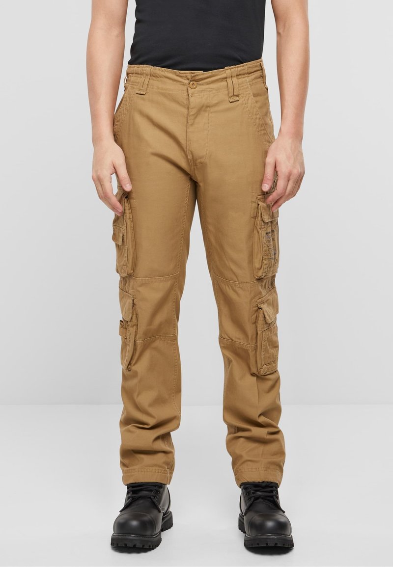 Homme portant un pantalon cargo beige avec plusieurs poches et des bottes noires, debout devant un fond gris clair uni.
