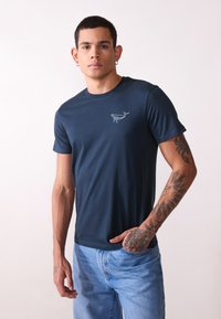T-shirt en coton bleu marine à manches courtes, col rond, avec un graphique de baleine blanche sur la poitrine gauche. Porté avec un jean bleu clair.