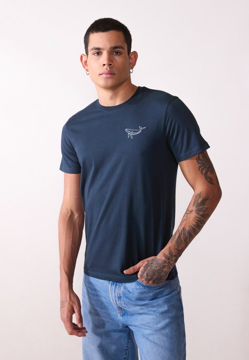 T-shirt en coton bleu marine à manches courtes, col rond, avec un graphique de baleine blanche sur la poitrine gauche. Porté avec un jean bleu clair.