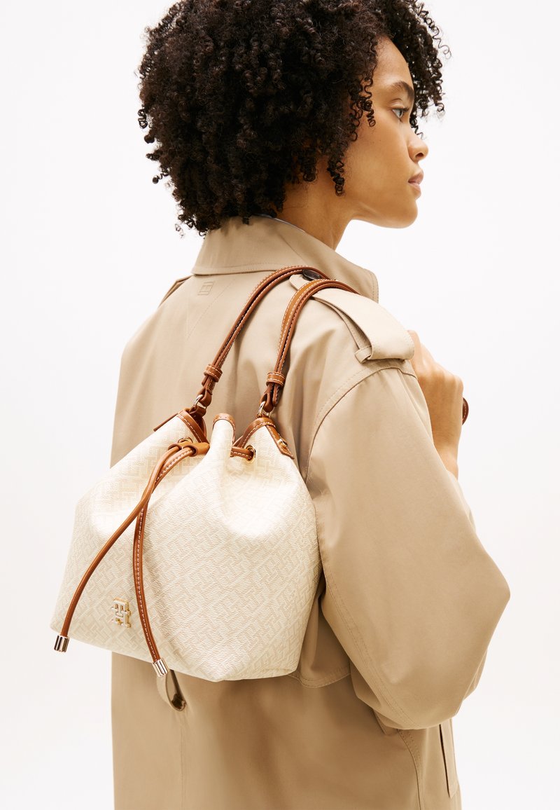 Γυναίκα με σγουρά μαλλιά, ντυμένη με μπεζ καμπαρντίνα, που κρατάει κρεμ και καφέ δερμάτινη τσάντα με κορδόνι στον ώμο.