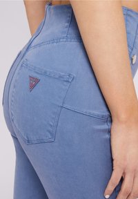 De hand van een vrouw rust op haar heup, gekleed in hoge blauwe spijkerbroeken met een driehoekig Guess-logo op de achterzak.