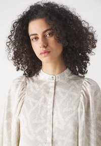 Blusa a maniche lunghe in tessuto bianco sporco con un motivo geometrico, dotata di maniche a sbuffo e colletto rotondo con chiusura a bottone.