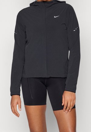 Person trägt schwarze Nike Kapuzenjacke und schwarze eng anliegende Radhosen, steht vor einem schlichten hellen Hintergrund.