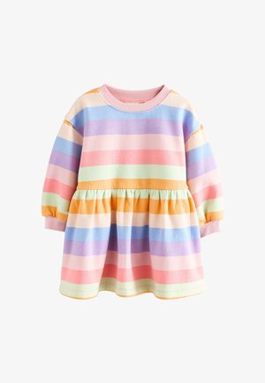 Jeune fille souriante, portant une robe à manches longues avec des rayures pastel horizontales, debout près d'un fauteuil beige texturé près d'une fenêtre.