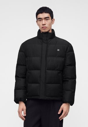 Jeune homme vêtu d'une veste matelassée noire à col haut, debout devant un fond clair uni, faisant face à l'objectif.