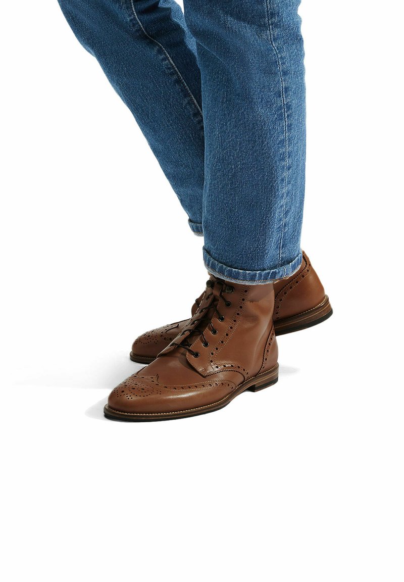 Stivali alla caviglia in pelle marrone con dettagli brogue, punta rotonda e lacci neri, abbinati a jeans blu con risvolto.