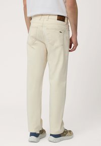 Pantalones de algodón beige con corte recto, que cuentan con dos bolsillos traseros y un parche de cuero en la cinturilla. Combinados con zapatillas grises.
