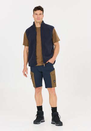 Mann trägt marineblauen Fleeceweste, braunes kurzärmeliges Hemd, zweifarbige Cargoshorts, schwarze Socken und Wanderschuhe, steht vor weißem Hintergrund.
