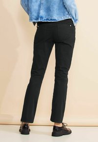 Svarta jeans med slim fit och standardfickor, i kombination med svarta läderskor. Bakgrunden är en neutral beige ton.