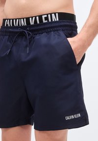 Μπλε σκούρο σορτς για κολύμβηση από ελαφρύ ύφασμα, με ρυθμιζόμενη μέση που διαθέτει ασπρόμαυρο λογότυπο και πλαϊνές τσέπες.