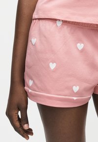 Gros plan d'une personne à la peau foncée portant un short rose avec des imprimés de cœurs blancs et un haut rose assorti, bras détendu le long du corps.
