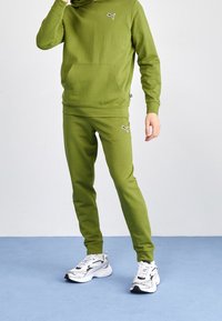 Olivgrön huvtröja med framficka och logotyp, matchad med olivgröna joggers. Bärs med vita och svarta sneakers på en blå yta.