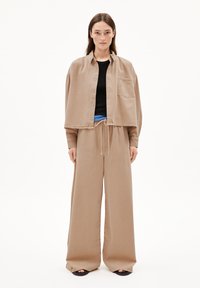Veste courte beige avec une poche sur un haut noir, associée à un pantalon beige ample. La tenue présente une taille à cordon et des jambes larges.