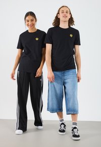 T-shirts en coton noir avec logo jaune, associés à un pantalon noir à rayures et à des culottes en denim. Les chaussures sont des baskets noires avec des accents blancs.