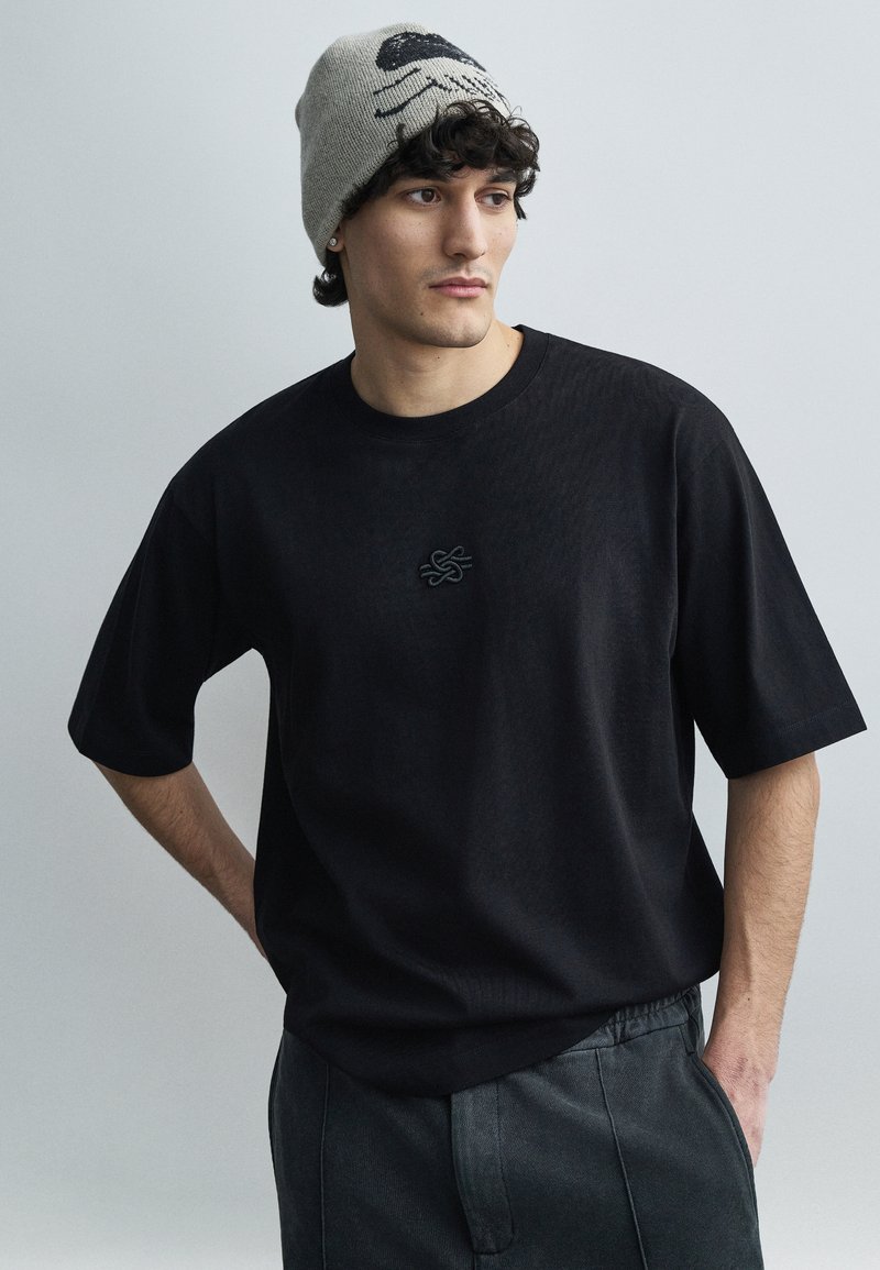 Black short-sleeve t-shirt with a textured finish and an embroidered emblem on the chest, paired with gray pants and a light knit beanie featuring a graphic.