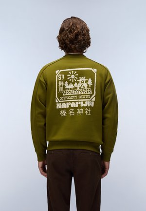 Personne aux cheveux bouclés porte un sweat-shirt vert olive avec un graphisme blanc représentant un soleil, des arbres, un bâtiment, des coordonnées et du texte dans le dos.