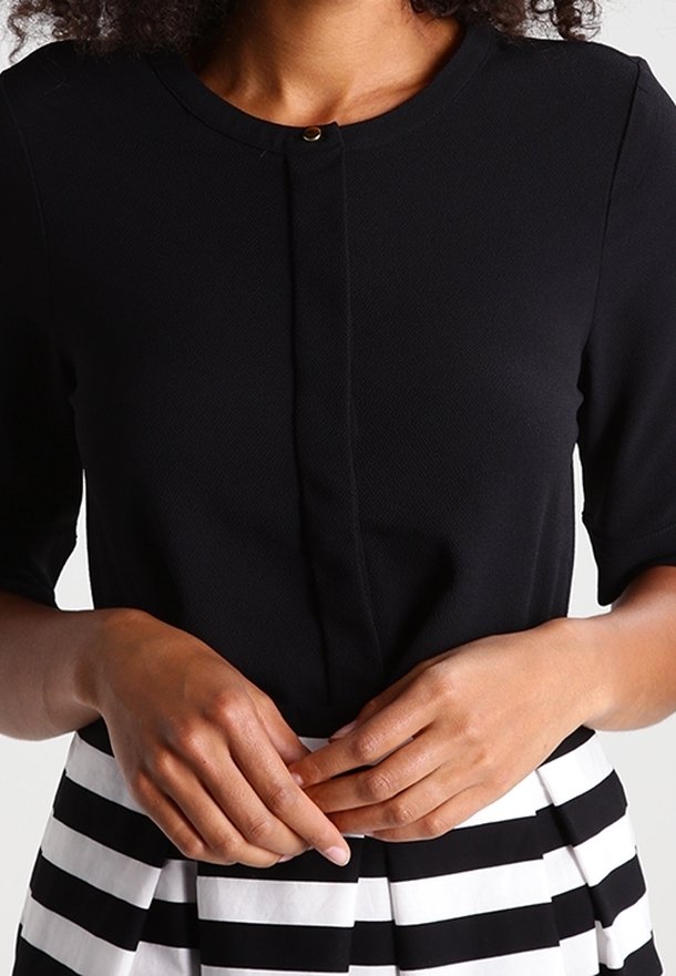 Femme portant un haut noir à col rond avec un petit bouton au niveau du cou et une jupe rayée noire et blanche à taille haute, les mains jointes.