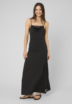 Femme souriante, portant une robe maxi noire sans manches avec une bordure en dentelle et des sandales noires, debout devant un fond gris uni.