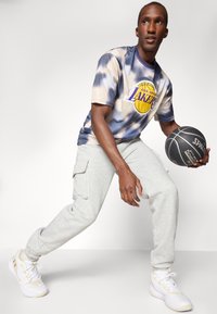 Grafisk t-shirt i blå og beige tie-dye med "Los Angeles Lakers" logo, kombineret med grå sweatpants og hvide sportsko, mens der holdes en sort basketball.