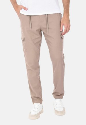 Uomo che indossa pantaloni cargo beige con tasche laterali, sneaker bianche e una camicia bianca, in piedi con una mano in tasca.