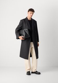 Mann in einem langen, dunklen karierten Mantel, schwarzem Hemd, hellfarbenen Hosen und schwarzen Schuhen, der eine große schwarze Leder-Clutch hält.