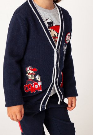 Marineblå cardigan med hvid kontrastkant, knaplukning og to lommer. Indeholder broderet "COACH" og figurgrafik. Blød strikket tekstur.