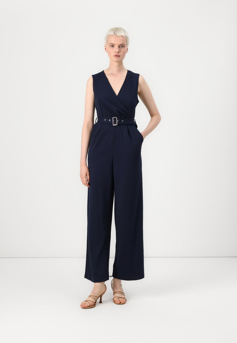 Tuta navy con un corpino aderente, scollatura a V profonda, gambe larghe e vita con cintura. Il tessuto appare liscio e strutturato. Tacchi a strappo completano il look.