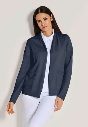 Vrouw met donker haar die een marineblauwe open vest draagt over een wit overhemd en witte broek, staand tegen een bleke achtergrond.