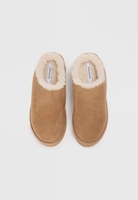 Tan suede slippers with a fluffy cream lining and rounded toe. Flat sole with minimal stitching detail, suitable for indoor use.