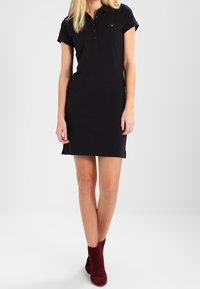 Robe polo noire à manches courtes, avec une patte de boutonnage à trois boutons et des fentes sur les côtés, associée à des bottines bordeaux. Texture de tissu lisse.