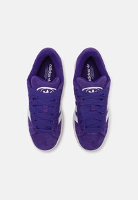 Fialové semišové tenisky s bílými akcenty, s texturovaným svrškem, kulatou špičkou a silnými tkaničkami. Logo je viditelné na vnitřní stélce.